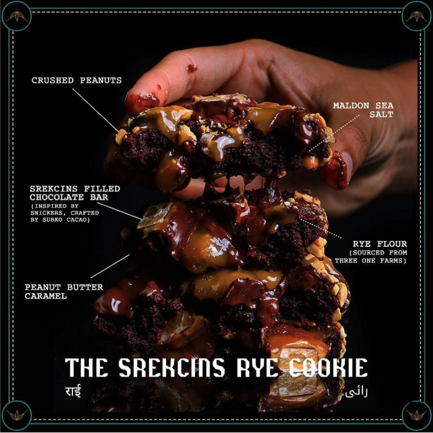Image showing a close-up of a dessert cookie with multiple labeled ingredients such as crushed peanuts, chocolate chunks, peanut butter caramel, Rye flour and sea salt, highlighting a product-focused food visual.